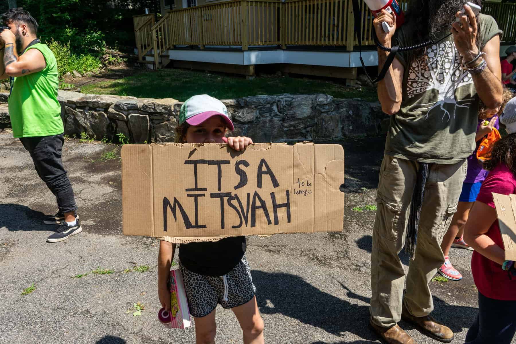 It's a happy girl holding a handmade cardboard sign that says "ITS A MITSVAH," likely at a Jewish camp or community event, surrounded by children and adults in casual outdoor attire.